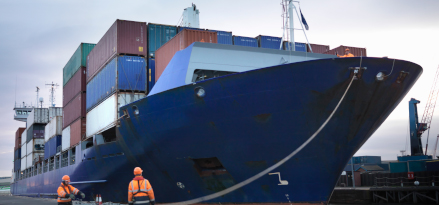 A blue cargo ship docked at a port loaded with shipping containers.